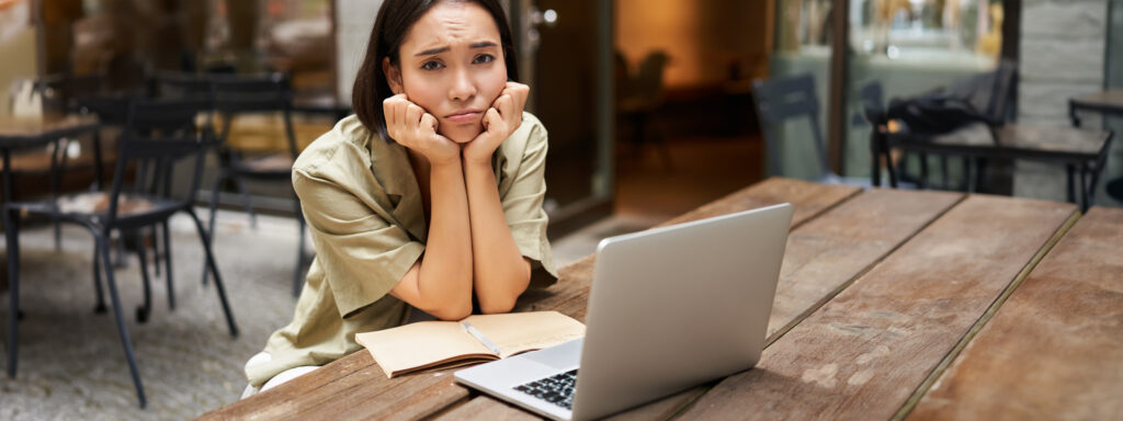 bored girl looks at her laptop, sits outdoors in cafe, listening boring online meeting, working and feeling sad